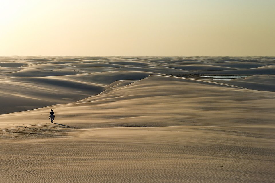 A small figure in silhouette walking across wide rippled pale-gold dunes in hazy evening light, a few more tiny figures far off