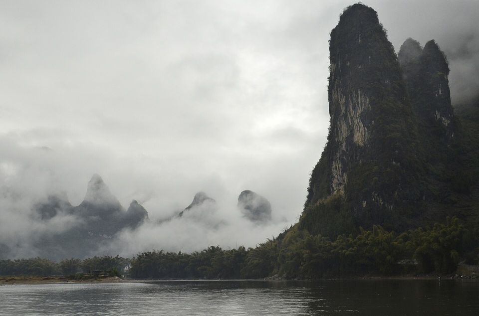 Dark limestone towers rising from a misty river, white cloud lying between the peaks and bamboo along the bank