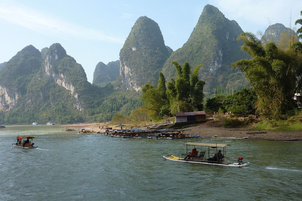 Two motor rafts with canopies on a green river below three steep limestone towers and bamboo, moored boats along a gravel landing