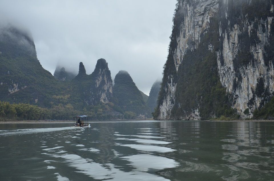 A small canopied raft on a grey-green river between steep limestone walls, low cloud on the peaks ahead