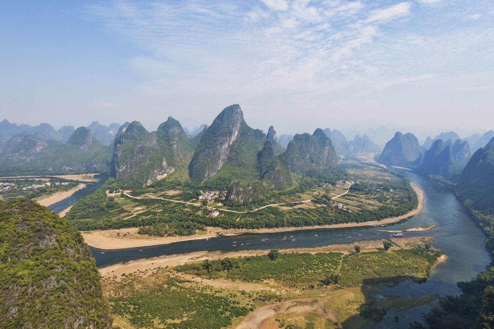 A blue-grey river bending across a green plain and pale sandbars, with rows of steep green limestone towers fading into haze