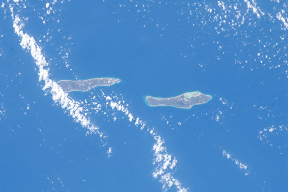 Two narrow, low islands ringed by pale reef lie in deep-blue ocean under lines of small white clouds
