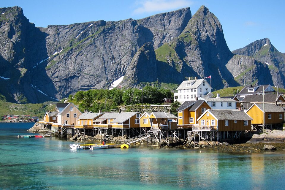 Ochre-yellow wooden cabins standing on stilts over clear turquoise water, sheer grey granite mountains rising behind