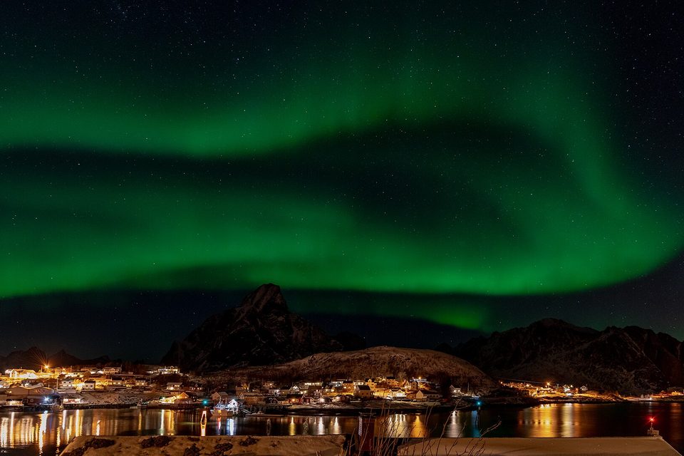 Broad green bands of aurora across a starry sky above a dark peak and the lit houses of a village reflected in the harbour