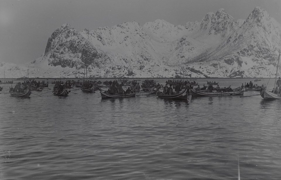 A black-and-white view of dozens of small open fishing boats crowded on calm water below a range of snow-covered mountains