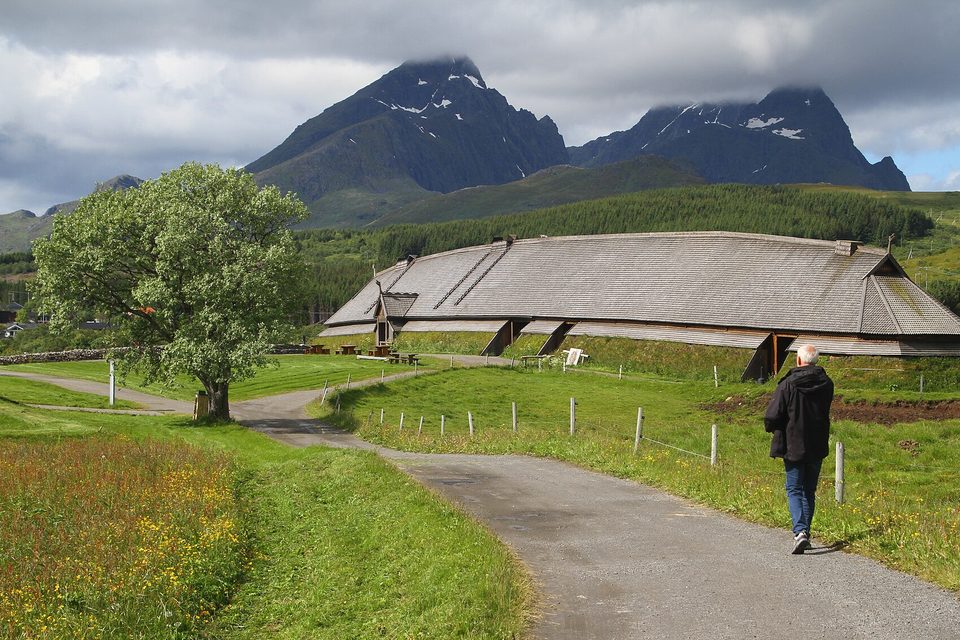 A long, low turf-green hall with a shingled roof curving along a hillside, a path and a tree in front, dark peaks in cloud behind
