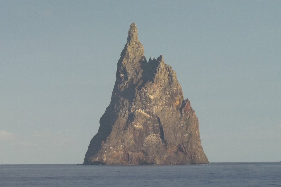A single sheer, jagged rock pinnacle rising straight out of a calm grey-blue sea in evening light