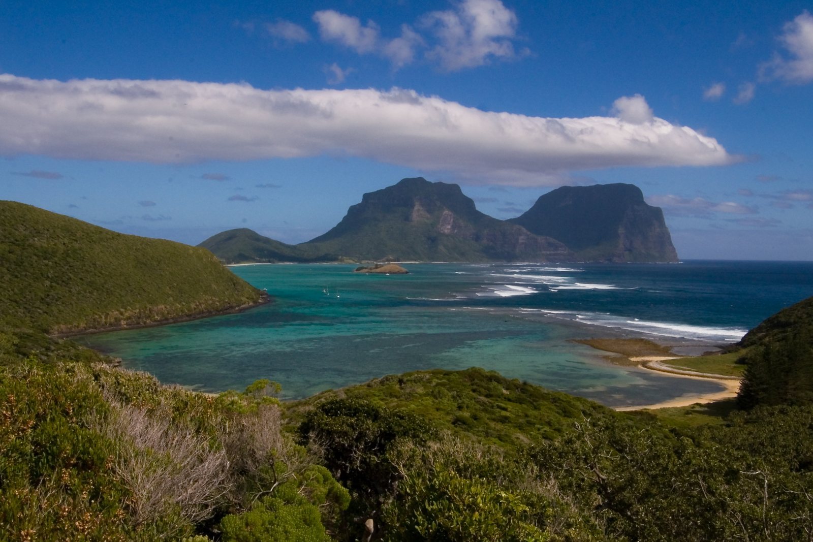 A low green crescent island enclosing a pale turquoise lagoon, two forested mountains rising at the far end under cloud