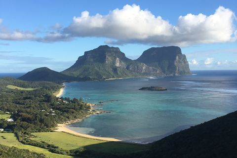 Two steep, flat-topped mountains rising above a turquoise lagoon and a curve of pale sand, with green hills and forest in the foreground and a cloud above the peaks
