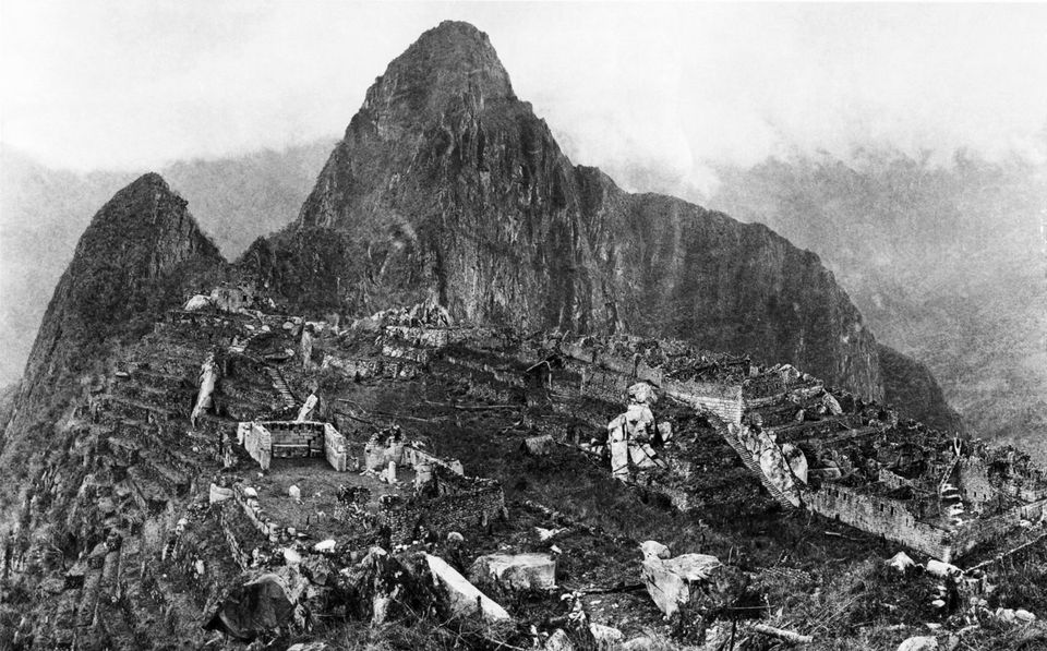 A black-and-white photograph of the ruins on a ridge below a steep peak, terraces on the left and a sacred hill on the right