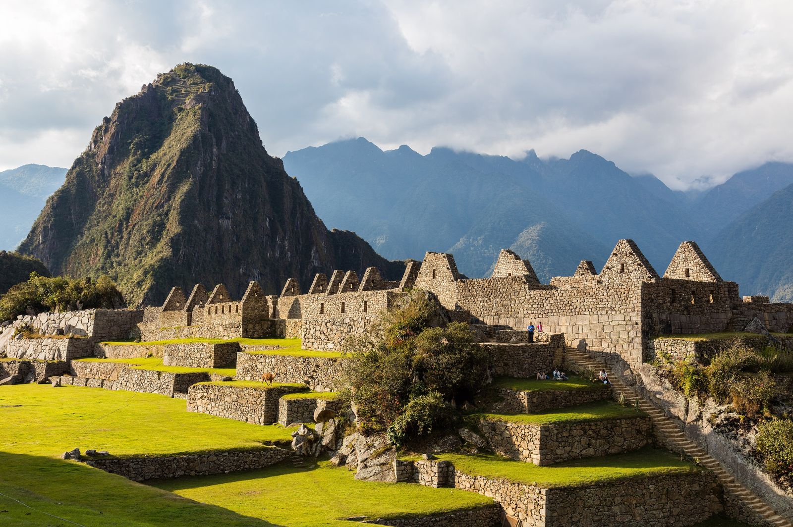 The Inca citadel of Machu Picchu in low golden light: green terraces and a row of gabled stone buildings below the steep peak of Huayna Picchu, cloud-shadowed mountains beyond
