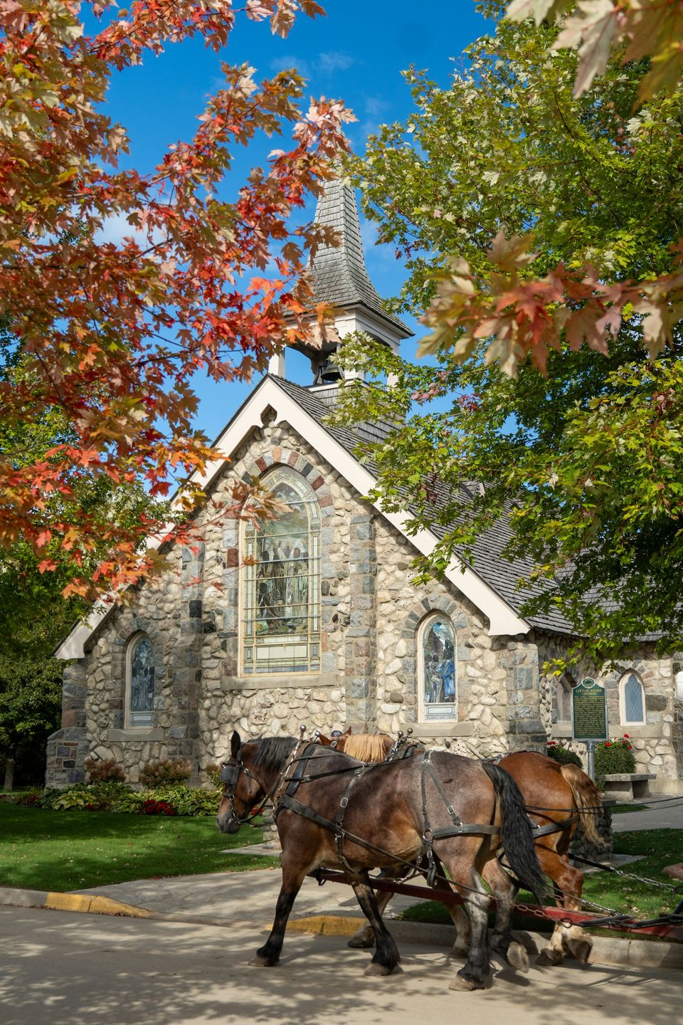Two draught horses pulling a carriage past a stone church among autumn trees
