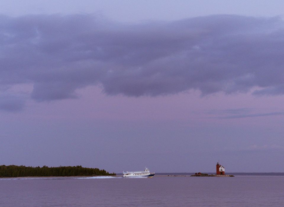 A passenger ferry crossing the channel past a red-and-white lighthouse and the wooded shore of an island