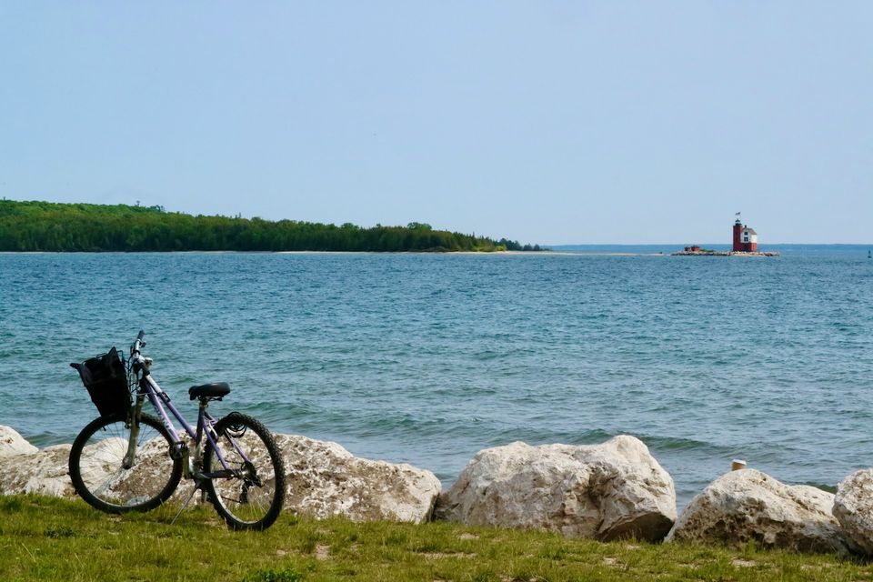A bicycle with a basket leaning on limestone rocks beside blue lake water, a red lighthouse far across the water