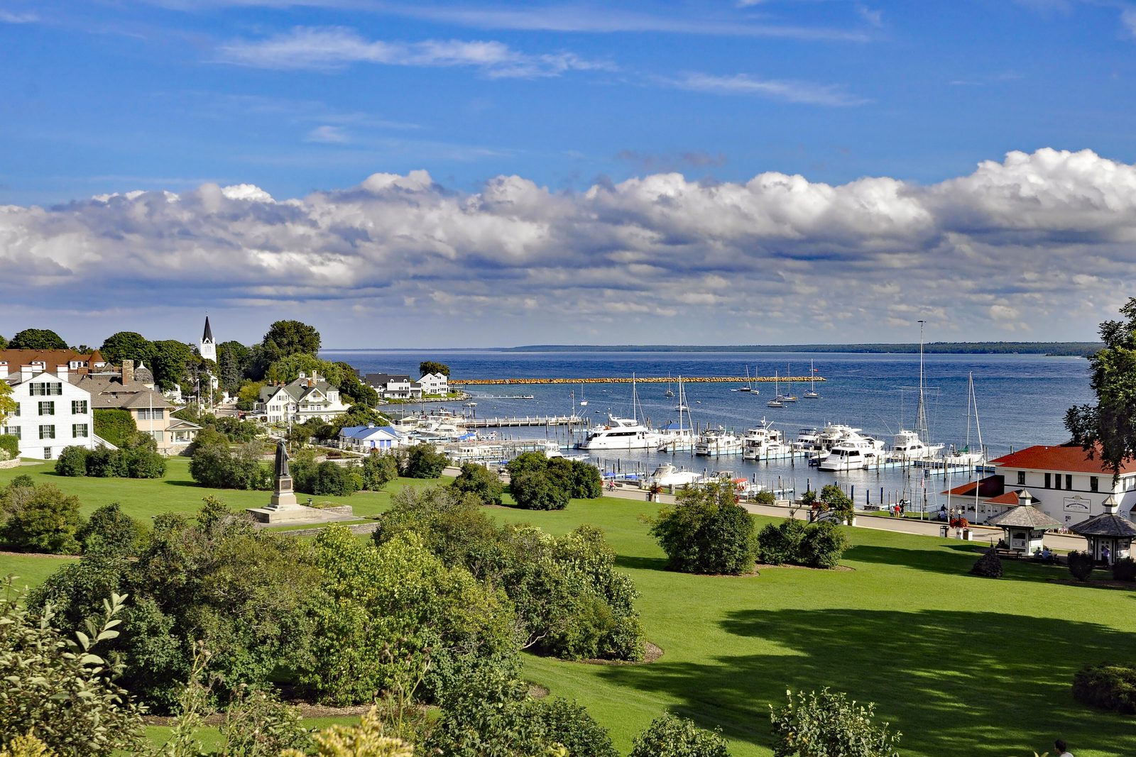 A green park and white houses sloping to a harbour full of moored boats on a wide blue lake under broken cloud