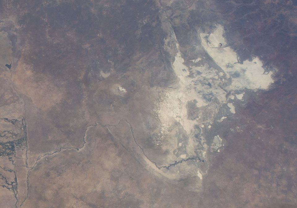 A view from orbit of brown savannah with a large irregular white salt pan and a thin winding river