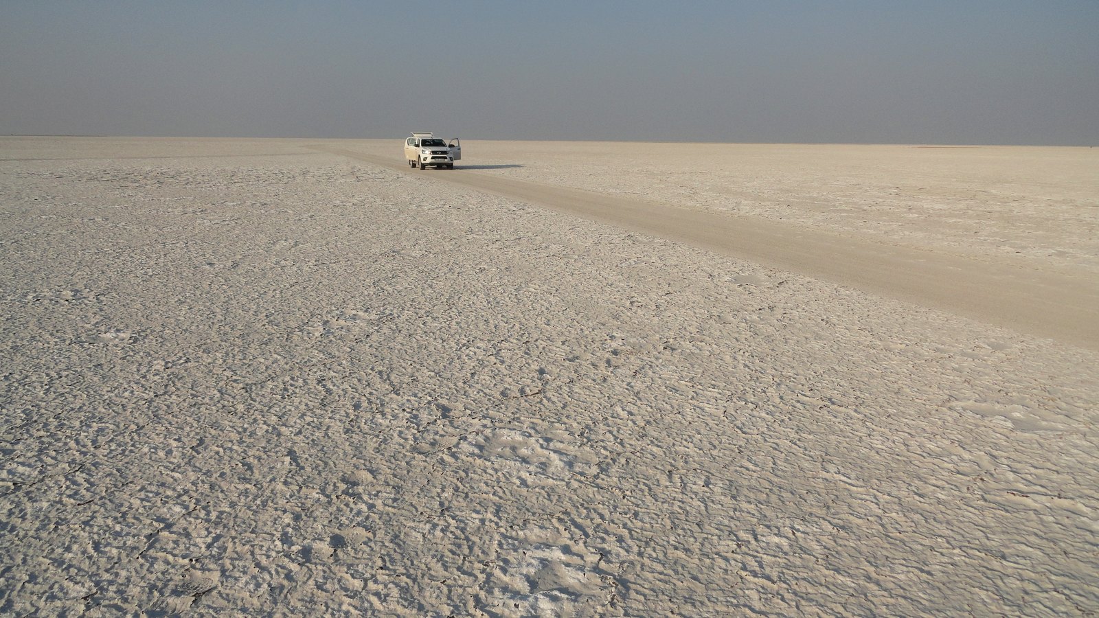 A flat white salt pan stretching to a hazy horizon, cracked crust in the foreground, a single white pickup truck parked far off beside a faint vehicle track