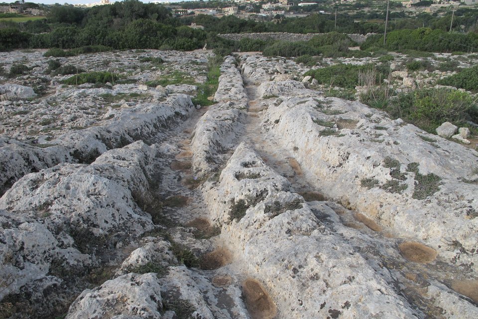 Pairs of deep parallel grooves cut into pale limestone running away across scrubby ground and crossing one another, with a dry-stone wall, trees and a village on the horizon