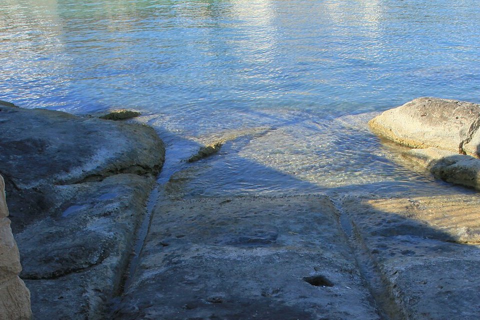 Two parallel grooves in a slab of dark rock running down into shallow, clear blue-green water, a pale rock ledge at the right