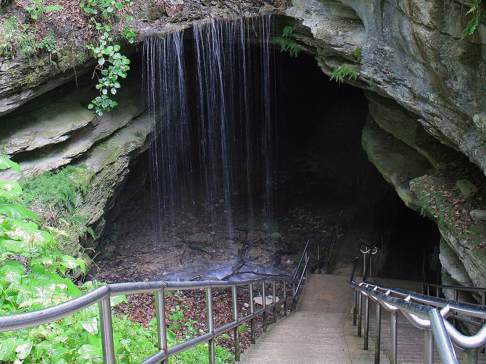 Stone steps with railings leading down into a dark cave mouth, a thin curtain of water falling from the ledge above, green ferns