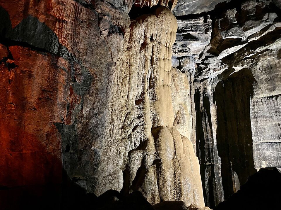A tall cream flowstone column against dark layered walls, warm light on the left