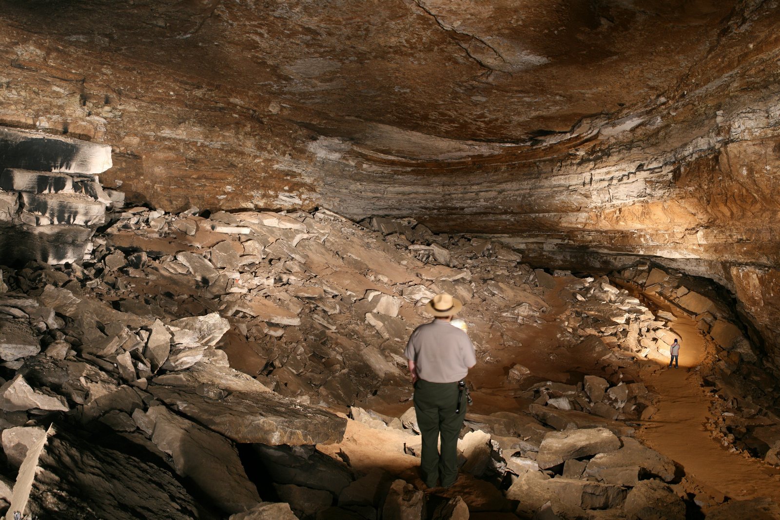 A park ranger in a flat hat stands on a rubble floor beneath a vast arched cave ceiling, a second figure tiny in the lit passage far behind