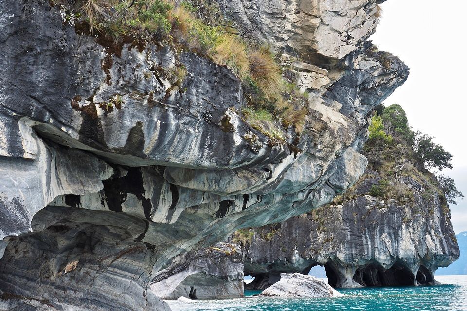 Grey and white banded marble cliffs undercut by smooth hollows above turquoise water, a rocky islet with trees behind and pale sky at the right