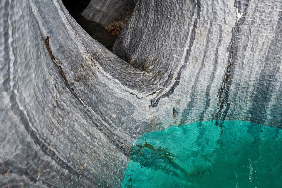 A curved wall of rippled grey and white banded marble dropping into a patch of bright turquoise water at the lower right