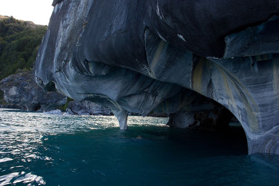 A huge overhang of smooth blue-grey marble with ochre streaks arching over dark green-blue water, forest on the shore at the left