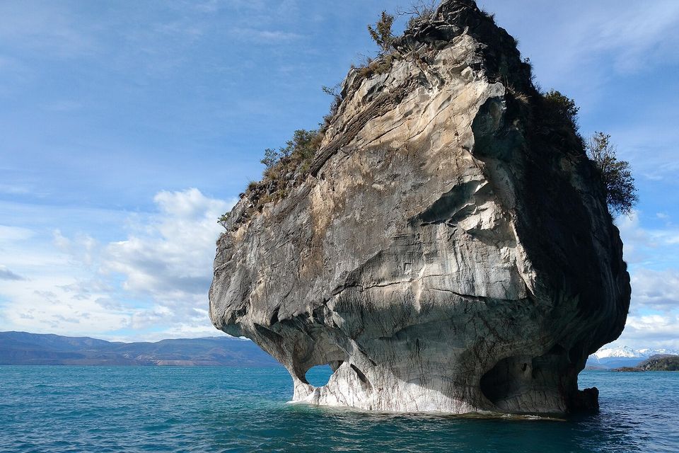 A large marble rock topped with a few trees standing alone in blue-green lake water, mountains with a little snow on the far shore