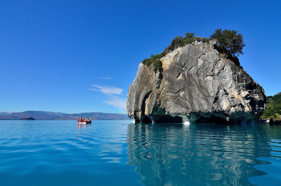 A rounded grey marble island topped with trees, reflected in deep blue water under a clear blue sky, a small boat far off at the left