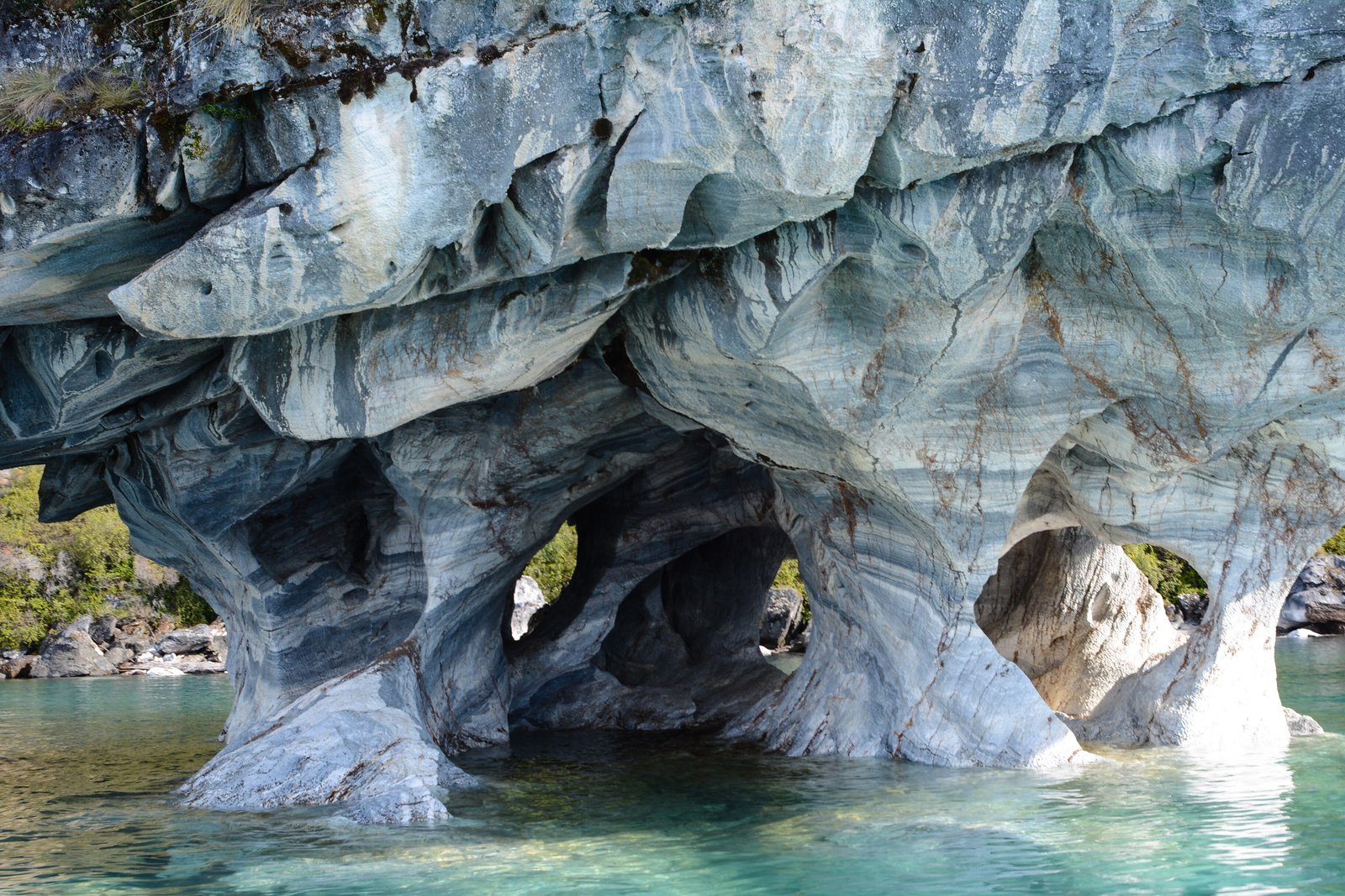 Pale grey and white banded marble hollowed into a row of arches and pillars standing in clear turquoise water, green forest on the shore behind