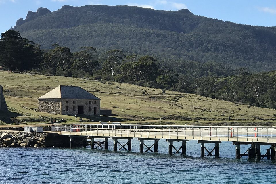 A sandstone building with a hipped roof on a grassy slope above a long jetty, below a forested mountain