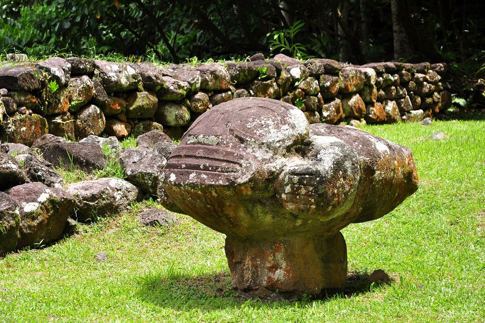 A weathered, lichen-spotted stone figure lying horizontally on a short pedestal on a lawn, a low wall of dark stones behind
