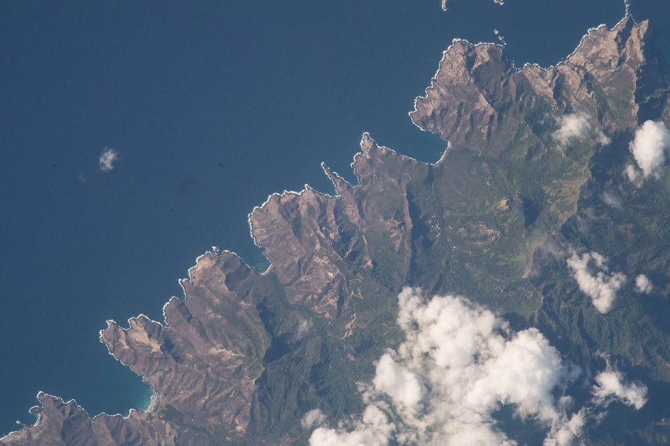 A photograph from orbit of brown, deeply ridged headlands dropping straight into dark blue ocean, a few white clouds over the land