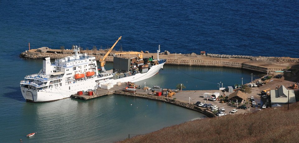 A white cargo-passenger ship with a yellow deck crane moored at a small concrete harbour below a dry hillside