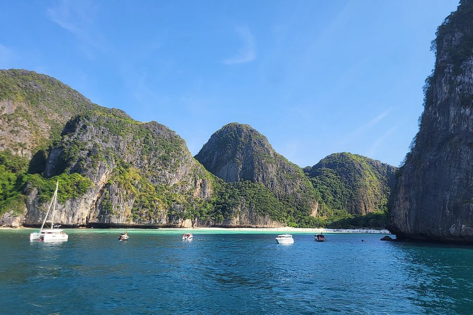 A bay ringed by tall limestone cliffs seen from the sea, a white beach along its foot and a few moored boats on blue water