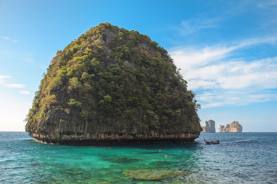 A dome-shaped rock island covered in scrub rising from turquoise water, two more rocks on the horizon and a longtail boat at the right