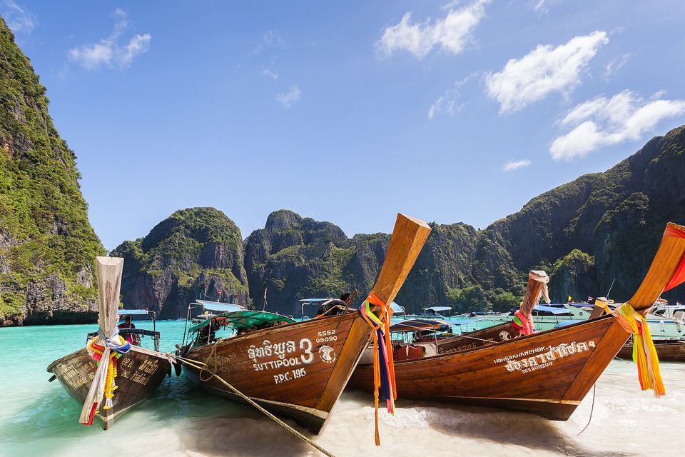 Three wooden longtail boats with bright ribbons pulled up on a white beach, turquoise water and a ring of limestone cliffs behind