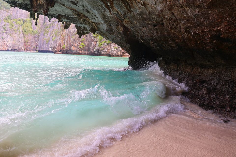 Turquoise water breaking in a curl of white foam onto pale sand beneath a dark rock overhang with hanging drip formations, grey-and-orange cliffs across the bay