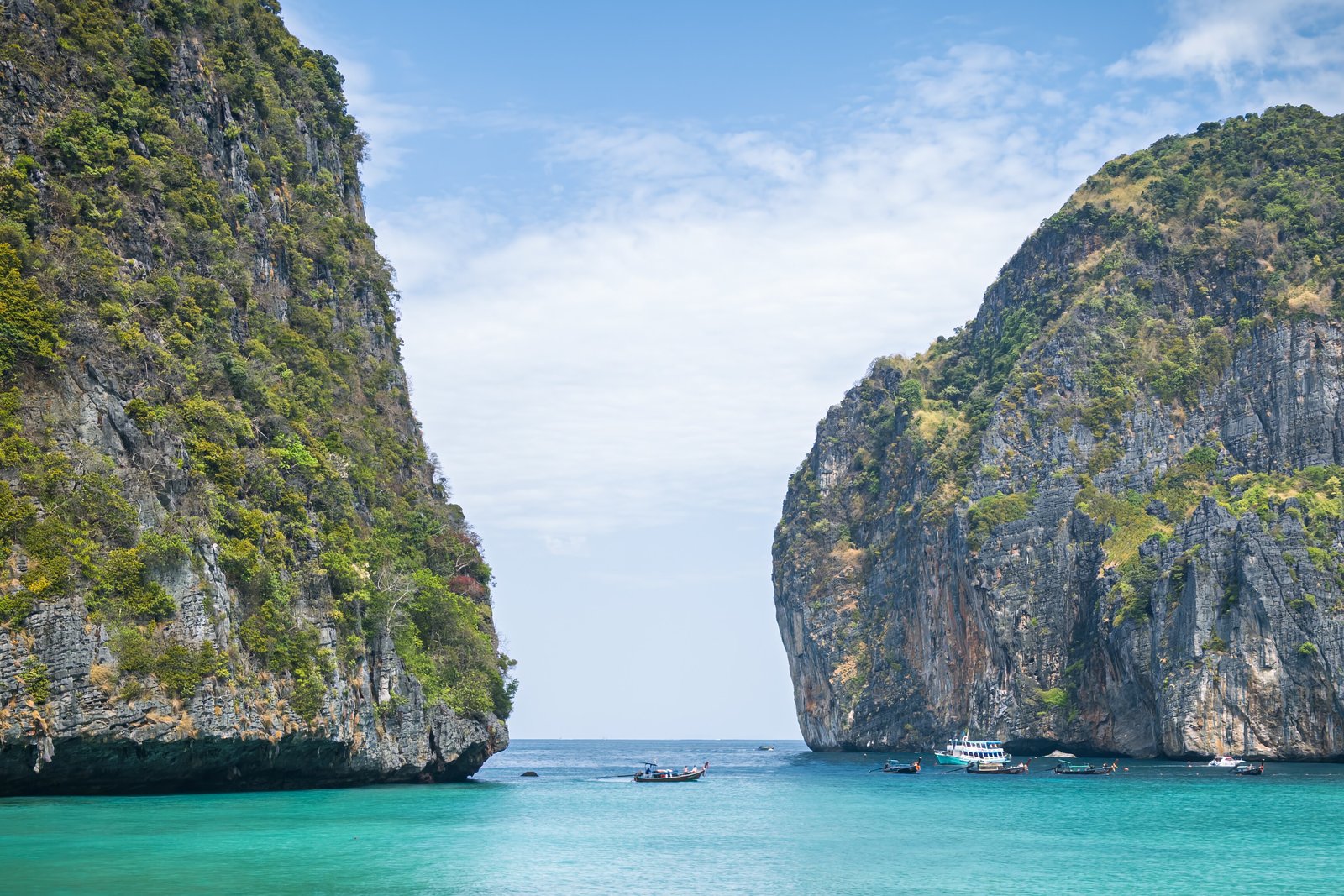 Two sheer limestone cliffs covered in green scrub frame a gap of pale sky above turquoise water, with small boats at the bay mouth