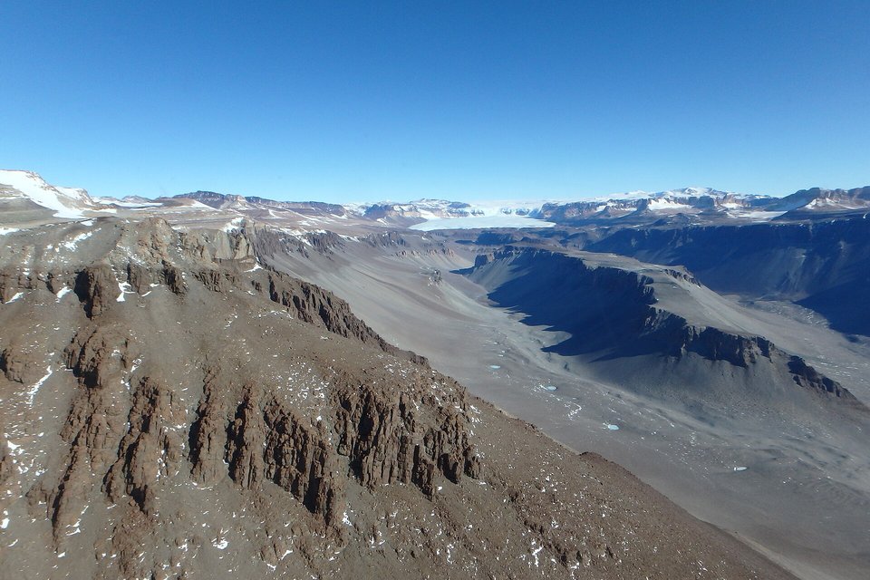 A wide ice-free valley of grey rubble running away between brown rock ridges under a clear deep blue sky, a pale icy expanse far off at its head