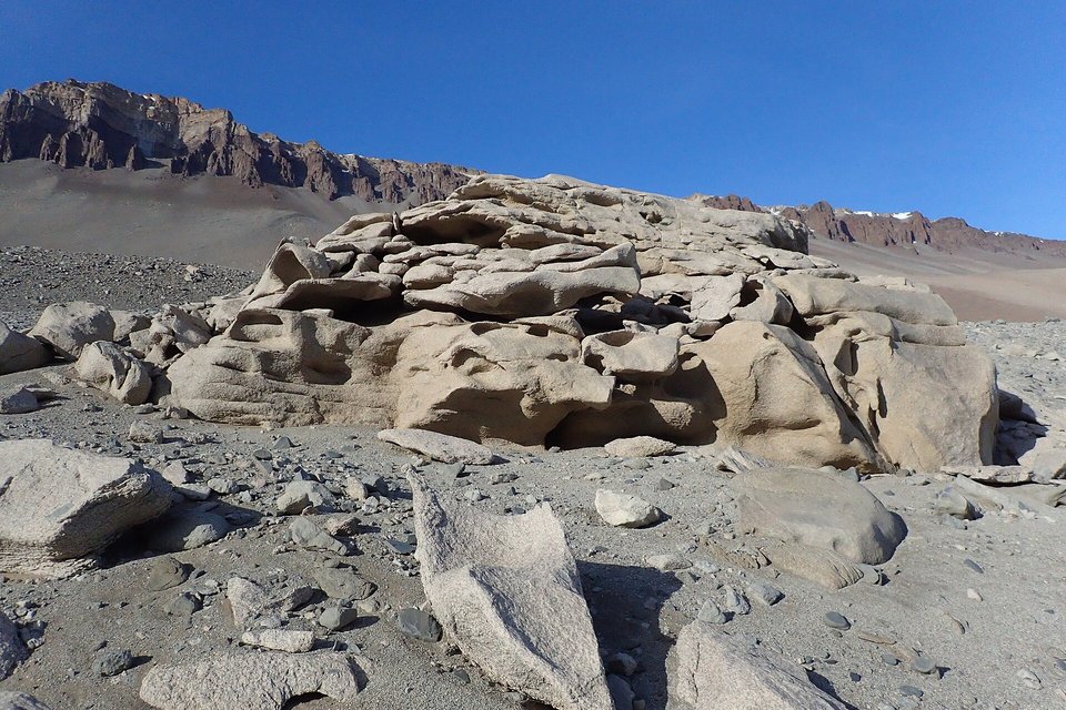 Pale wind-sculpted boulders on a gravel valley floor, a dark rock ridge and a deep blue sky behind