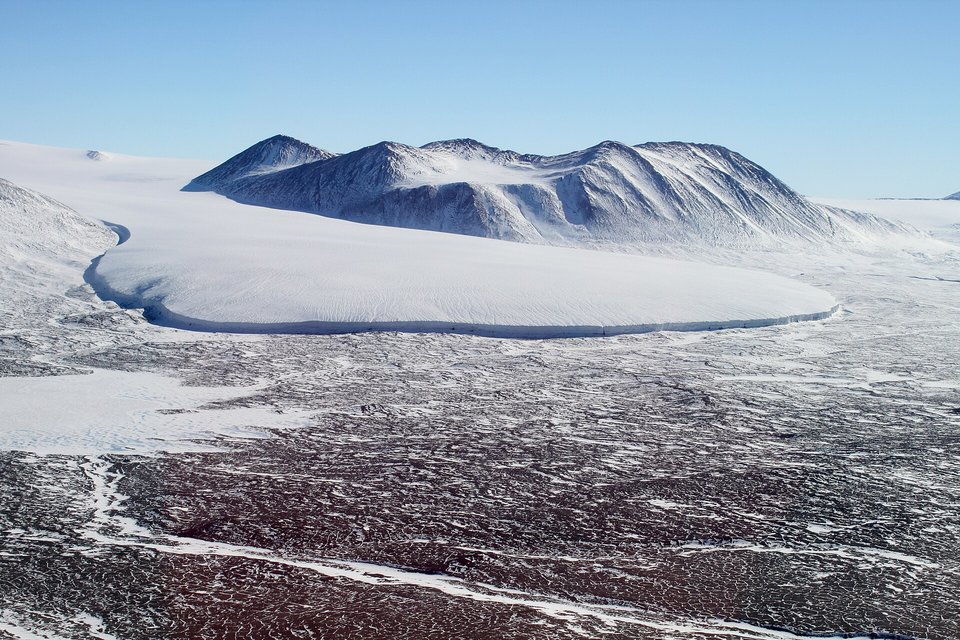 A broad white glacier tongue with a sharp rounded edge ending on a dark valley floor, snowy mountains behind it