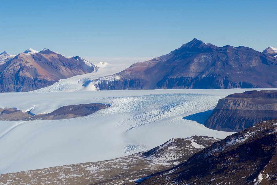 A broad white glacier flowing between brown mountain ridges under a clear sky