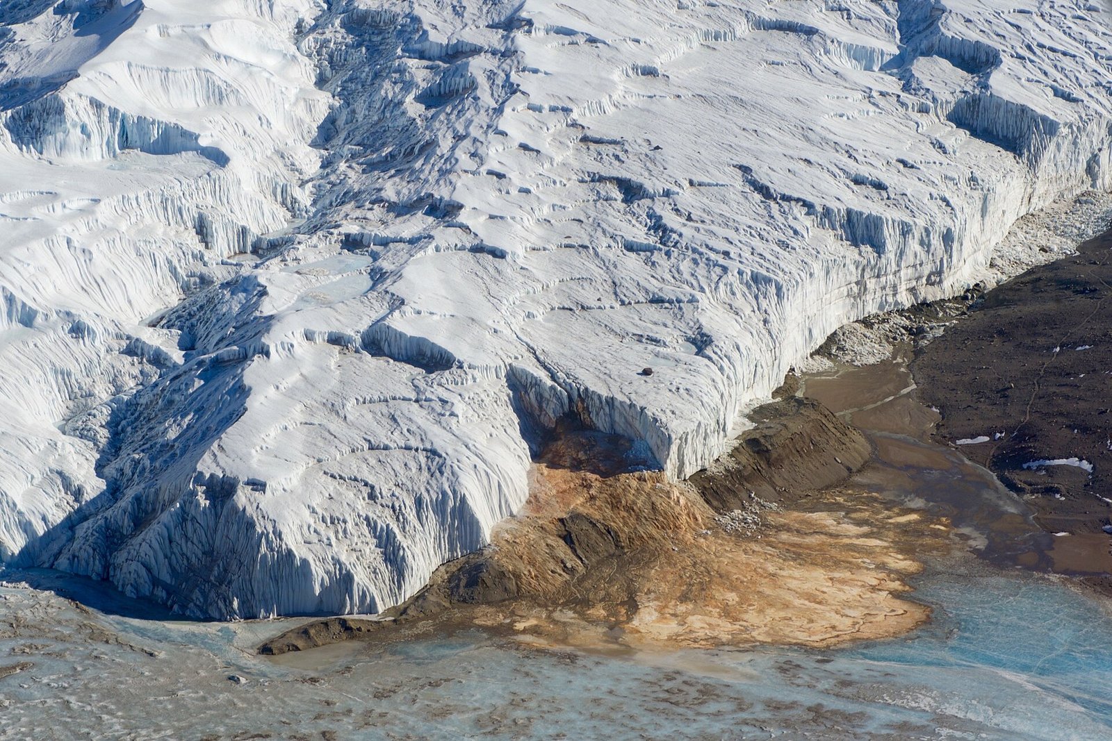 A wall of white glacier ice meeting a bare valley floor, a rust-orange stain of iron-rich brine spilling from its foot across the ground toward a frozen lake
