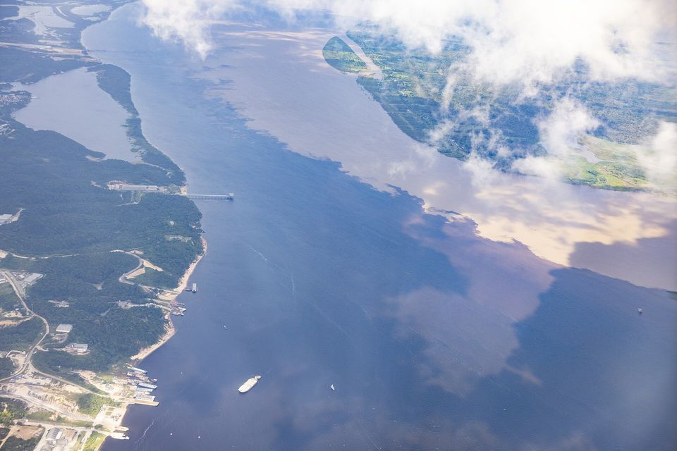 A wide river seen from a plane window, dark water and pale brown water meeting along a curving line, with forest and a small port on the left bank and cloud at the top