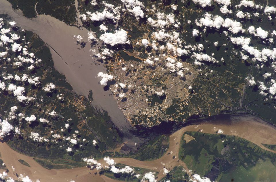 A satellite view of dark green forest and a grey city beside a wide silvery river that turns dark where it meets a tan river curving across the bottom, with scattered white clouds
