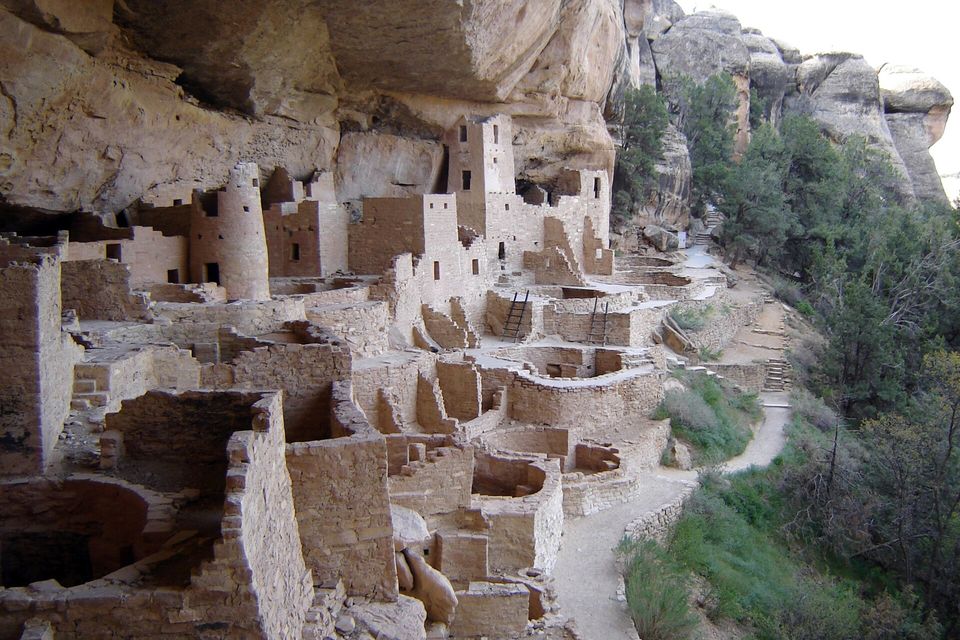 Dozens of stone rooms, round walled pits and towers filling a long alcove below a cliff, with a path beside them