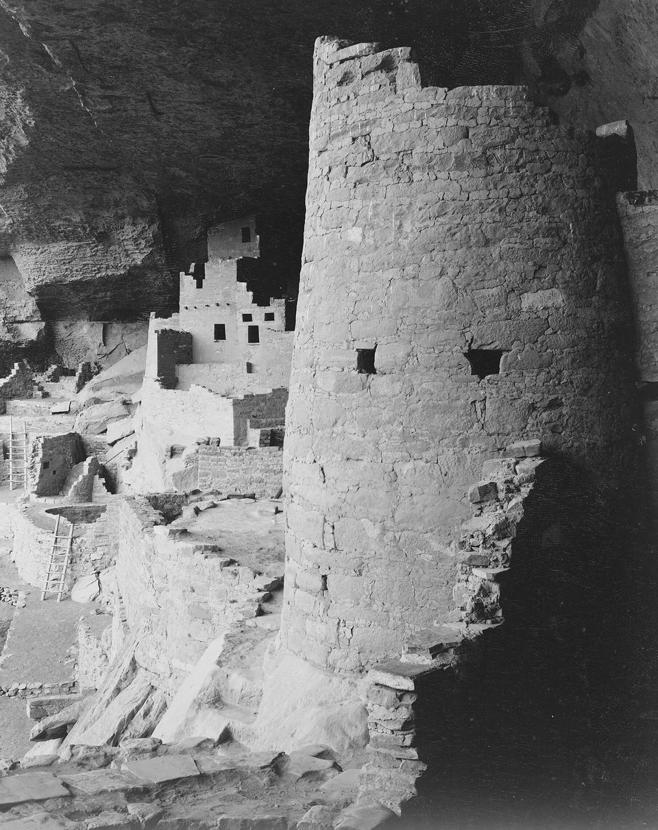 Black-and-white photograph of a round stone tower in front of stacked rooms under a cliff overhang